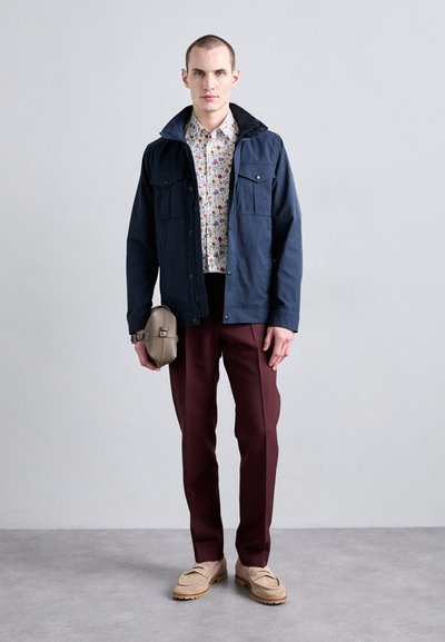 Jachetă bleumarin cu buzunare cu nasturi peste o cămașă albă cu flori, combinată cu pantaloni burgundy și mocasini bej. O geantă mică bej în mână.