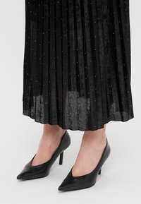 Sorte højhælede sko med spids tå, lavet af glat læder. Båret med en plisseret, glitrende sort nederdel med lodrette linjer.