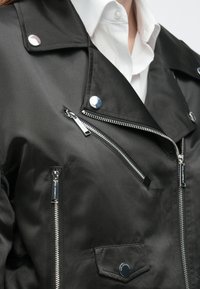 Čierna saténová motorkárska bunda so striebornými kovaniami, viacerými vreckami na zips a golierom so zapínaním na cvok, s hladkým povrchom.