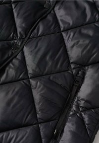 Veste matelassée noire avec finition brillante, boutons-pression et une poche verticale zippée avec un tirette en tissu.