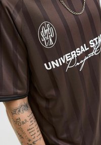 Brunstribet skjorte med hvid tekst "UNIVERSAL STATE", cirkulært "SOFICK" logo, sølvkæde halskæde og tatoveret underarm.