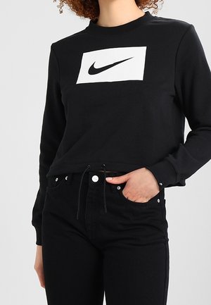 Personne portant un sweat-shirt Nike noir à manches longues avec logo blanc et pantalon noir, une main dans la poche, devant un fond gris.