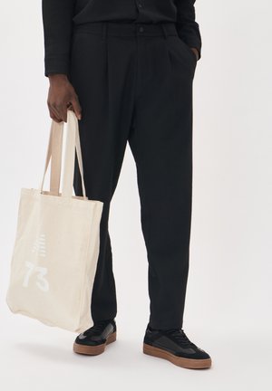 Pantaloni neri in tessuto strutturato con pieghe frontali, abbinati a sneakers nere con suole beige. Tiene una borsa a tracolla beige con grafiche bianche.