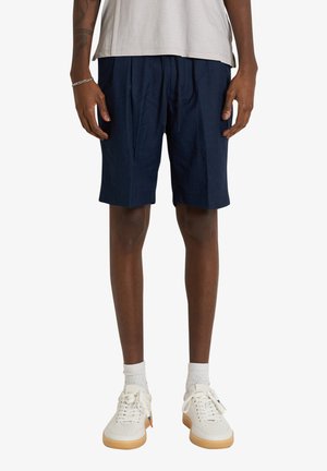 Uomo che indossa pantaloncini blu navy, sneakers bianche con suole in gomma, calzini bianchi e una camicia grigio chiaro su sfondo bianco.