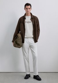 Camisa marrón de botones sobre una camiseta de color crema claro, pantalones blancos y zapatos negros. Lleva un bolso verde oliva. Diseño simple y colores apagados.