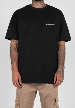 Mand iført sort t-shirt med hvid "MJGONZALES" tekst på brystet og beige cargobukser, der viser tatoverede arme og hænder med armbånd og ring.