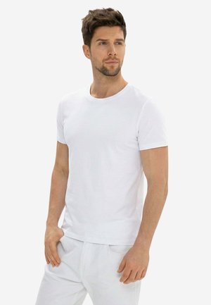 Uomo con capelli castani corti che indossa una maglietta bianca semplice a maniche corte e pantaloni bianchi, guarda di lato con una mano in tasca.