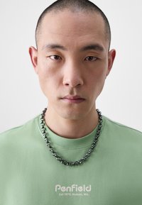 Zöld pamut póló "Penfield" logóval, vastag fém láncnyaklánccal kombinálva. A modellen kopasz fej és semleges arckifejezés látható.