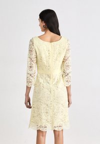 Robe en dentelle jaune doux, dotée de manches trois-quarts, d'un col rond et de détails à ourlet festonné. Fermeture éclair au dos.