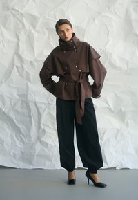 Nő barna, övvel díszített, magas galléros dzsekit visel, fekete, laza nadrágban, egy gyűrött fehér háttér előtt áll.