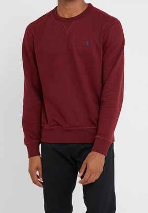 Mann trägt einen burgunderfarbenen Langarm-Pullover mit kleinem marineblauem Logo auf der linken Brustseite und schwarze Hose vor neutralem Hintergrund.