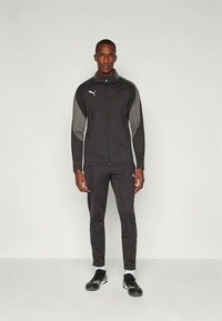 Melna sporta treniņtērps ar ar rāvējslēdzēju aizveramu jaku, kurai ir pelēki akcenti, un atbilstošām biksēm. Materiāls izskatās viegls un pieguļošs. Melni sporta apavi.