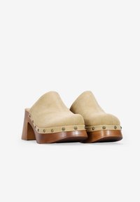 Zuecos de ante beige con punta redondeada, tacón de madera elevado y tachuelas de metal a lo largo de la base para añadir detalle. Textura suave con un diseño minimalista.