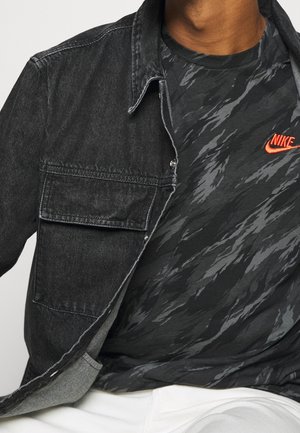 Mężczyzna w czarnej dżinsowej kurtce, na czarno-szarej moro koszulce Nike z pomarańczowym logo i białych spodniach, skróconych na szyi i talii.