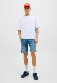 Witte oversized t-shirt, blauwe denim shorts met opgerolde zoom, marineblauwe sneakers, rode pet, effen witte sokken, minimale slijtage op de shorts.