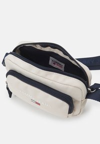 Borsa a tracolla compatta in tessuto beige con dettagli blu navy, dotata di due scomparti con cerniera e una toppa con logo. Tracolla regolabile inclusa.
