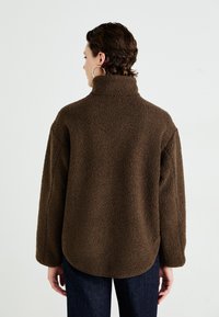 Person wearing a brown high-collar fleece jacket with long sleeves and dark pants, facing away against a plain white background.