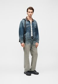 Τζιν μπουφάν με καφέ γιακά, λευκό αμάνικο τοπ, γκρι παντελόνι φαρδιάς γραμμής και μαύρα αθλητικά παπούτσια· το μοντέλο κρατά μια μαύρη τσάντα, στέκεται μπροστά σε λευκό φόντο.