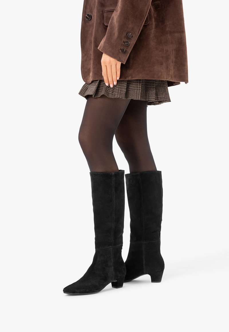 Personne portant des bottes en daim noir montant jusqu'aux genoux, des collants noirs transparents, une jupe plissée à carreaux et une veste en daim marron.