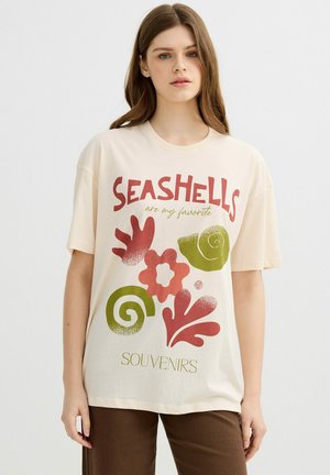 Joven mujer que lleva una camiseta beige con diseños de conchas marinas rojas y verdes y el texto "SEASHELLS son mis recuerdos favoritos" posando frente a un fondo blanco.