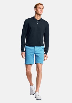 Marineblauw poloshirt met lange mouwen, lichtblauwe korte broek en witte sneakers. Het poloshirt heeft een kraag en knopen; de korte broek reikt tot de knieën.