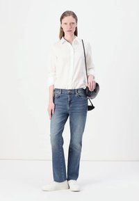 Camicia bianca a bottoni, jeans blu dritti, sneaker bianche, borsa a tracolla nera, in piedi contro uno sfondo neutro.