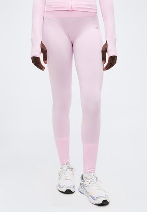 Person som har på seg blekrosa tettsittende leggings og matchende jakke, står med hvite og lilla treningssko på hvit bakgrunn.