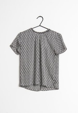 Musta-valkoinen lyhythihainen pusero geometrisella kuviolla, roikkuu mustassa henkariissa valkoista taustaa vasten.