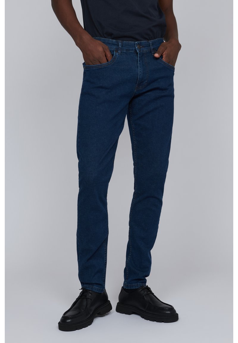 Vaqueros de mezclilla azul oscuro con un corte recto, que cuentan con bolsillos laterales, presillas para el cinturón y un diseño clásico de cinco bolsillos. Combinados con zapatos negros.