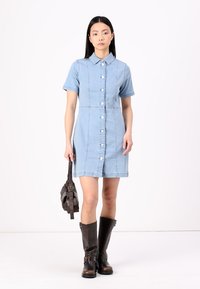 Svetlomodré denim šaty s krátkymi rukávmi, predným zapínaním na gombíky, švovými detailami a priliehavým strihom. V kombinácii s hnedými čižmami po kolená.
