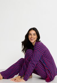 Pajama set s uzorkom pruga koje uključuju uske ljubičaste i ružičaste pruge, dugih rukava i opuštenog kroja, s košuljom na dugmad i širokim hlačama.