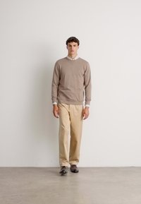 Homme marchant à l'intérieur portant un pull beige par-dessus une chemise blanche, un pantalon crème et des chaussures brunes devant un mur blanc uni.