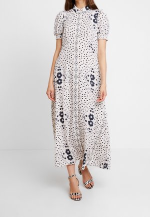 Femme vêtue d'une robe midi blanche à pois noirs, motifs floraux et diamants, manches bouffantes, et talons ouverts à rayures zèbre.