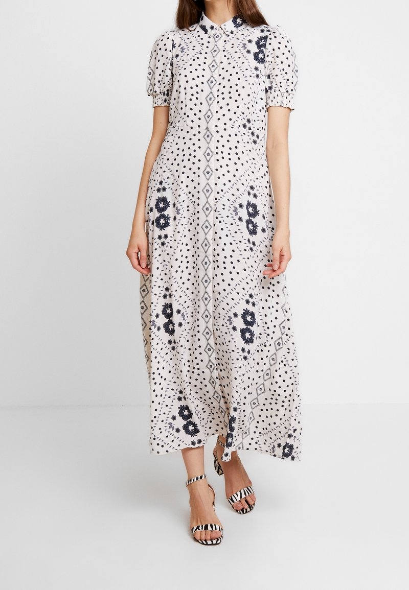 Femme vêtue d'une robe midi blanche à pois noirs, motifs floraux et diamants, manches bouffantes, et talons ouverts à rayures zèbre.
