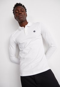 Garā piedurkņu balts polo krekls ar klasisku apkakli, uz krūtīm ir mazs tumši zils logo, apvienots ar tumšiem biksēm.