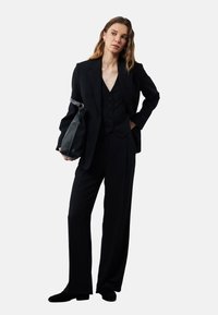 Zwart op maat gemaakt pak met een double-breasted blazer, een vest met knopen en wijde broekspijpen, gecombineerd met een zwarte handtas.