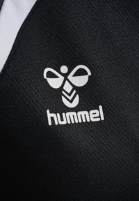 Wit Hummel-logo met gestileerde bij boven de tekst op een gestructureerde zwarte stof, onderdeel van een zwart-wit kledingstuk.