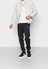 Osoba w jasnobeżowej kurtce, białym t-shircie, czarnych dresowych spodniach z niebieskimi logo oraz czarnych sneakersach z białymi podeszwami i logo Swoosh.