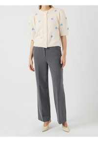 Crème-kleurige gebreide cardigan met blauwe bloemenborduursels, korte mouwen en knopen, gecombineerd met grijze op maat gemaakte broek en beige hakken.