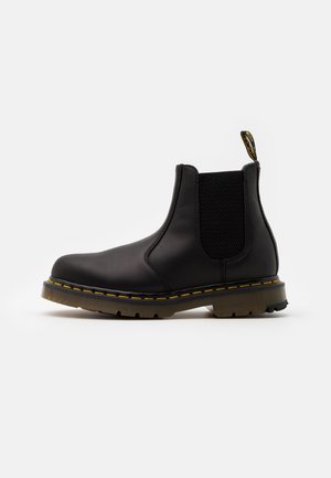 Bottines Chelsea en cuir noir avec des panneaux élastiques sur les côtés, des coutures jaunes et une semelle en caoutchouc. Comprend une languette pour un enfilage facile.