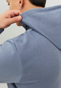 Niebieska bluza z miękkiego materiału, z kapturem na sznurku i żebrowanym wykończeniem. Dłoń chwytająca kaptur, ukazująca detale szwu.