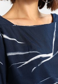 Maglietta blu navy con scollatura ampia, decorata con linee astratte bianche. Il tessuto appare morbido e dalla texture interessante, con una vestibilità comoda.