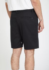 Man draagt zwarte knielange korte broek met achterzakken en kleine logotags, staand tegen een effen witte achtergrond.