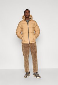 Bež jakna s punjenjem i kapuljačom, s prednjim patentnim zatvaračem i elastičnim manžetama, u kombinaciji s smeškim končanim hlačama i crnim tenisicama s bijelim detaljima.