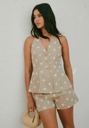 Femme aux cheveux foncés portant un top beige sans manches boutonné et un short assorti avec des imprimés cachemire blancs, tenant un petit sac à main tressé.