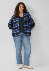 Frau mit lockigem Haar, die ein blaues karriertes Hemd über einem schwarzen Oberteil, hellblaue Jeans und weiße Sneakers trägt, steht vor einem grauen Hintergrund.