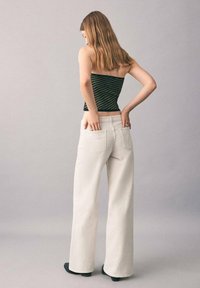Top ajustado a rayas negras y verdes; jeans de pierna ancha blancos con bolsillos traseros; textura suave; modelo visto desde atrás.
