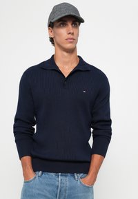 Jonge man in een grijze pet, marineblauwe geribbelde trui met kraag en lichtblauwe jeans, staand met handen in zijn zakken tegen een witte achtergrond.