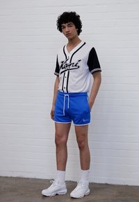 Hvit og svart kortermet jersey med blå shorts, har en logo og kontrastdetaljer, kombinert med hvite joggesko og sokker.
