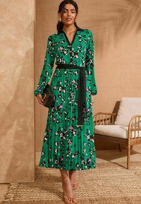 Abito verde a fiori con pieghe, maniche lunghe, colletto nero e cintura. Presenta motivi multicolor ed è abbinato a una borsa nera a mano e sandali color sabbia.